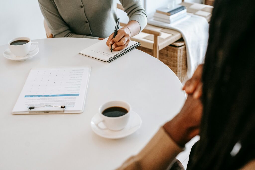 pexels-photo-5699480-5699480 From above crop multiracial female interviewer or psychologist in formal wear asking questions and taking notes in planner while talking to black man and sitting together at table with coffee